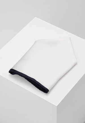 Accessorio tessile bianco con superficie testurizzata e un bordo in navy scuro lungo il fondo, piegato su una piattaforma bianca.