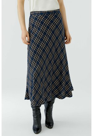 Vrouw draagt een blauw-zwart geruite midi-rok met zwarte leren laarzen en een wit topje, staand op een lichte achtergrond.
