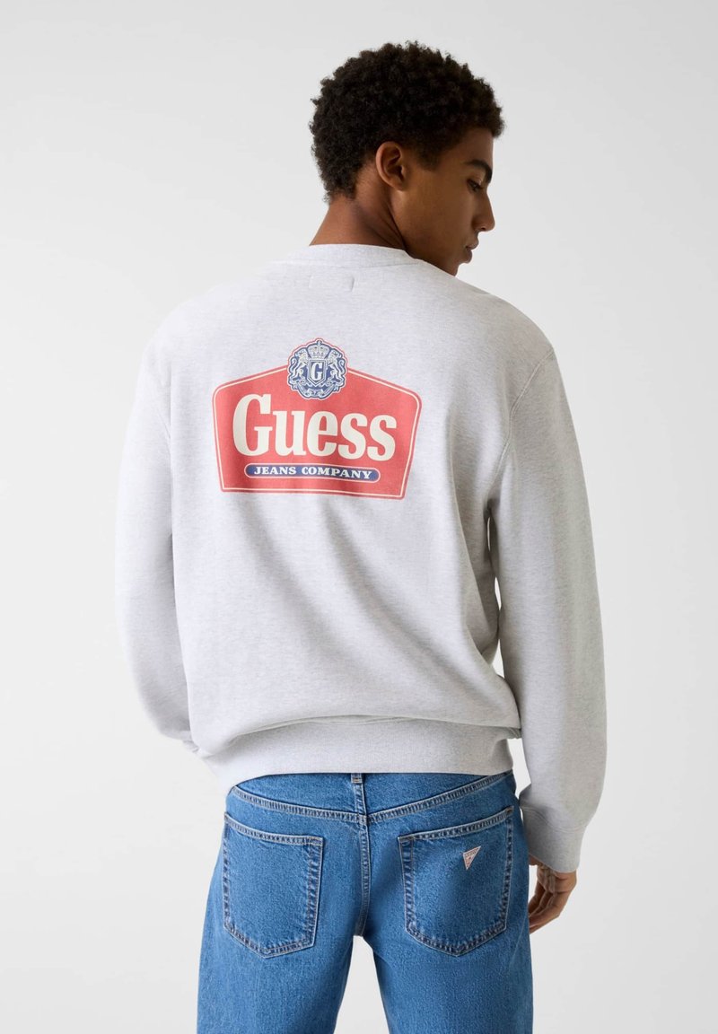 Felpa grigia con il logo rosso e blu "Guess Jeans Company" sul retro, indossata con jeans di denim blu, dotati di tasche laterali.