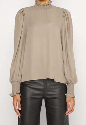 Beżowa bluzka z długim rękawem, z falbanką przy wysokim kołnierzu, bufkami na ramionach oraz marszczonymi mankietami, zestawiona z czarnymi skórzanymi spodniami, na stojącej osobie.