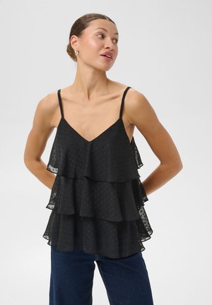 Une femme pose les mains sur les hanches, vêtue d’un caraco noir à volants superposés et d’un jean bleu foncé, sur un fond clair et uni.