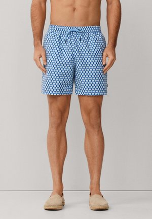 Man draagt blauwe zwemshorts met patroon en beige instapespadrilles, staand tegen een effen lichtgrijze achtergrond.