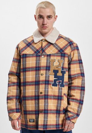 Tan plaid shirt jacket with a soft, cream-colored collar. Features blue and brown embroidered letters on the left chest. Buttons down the front.
