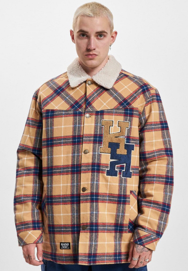 Tan plaid shirt jacket with a soft, cream-colored collar. Features blue and brown embroidered letters on the left chest. Buttons down the front.