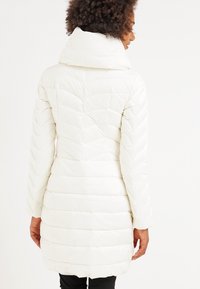 Vit puffjacka med hög krage, som har en kviltad design och långa ärmar. Baksidan visar vertikal söm och en medellång längd.