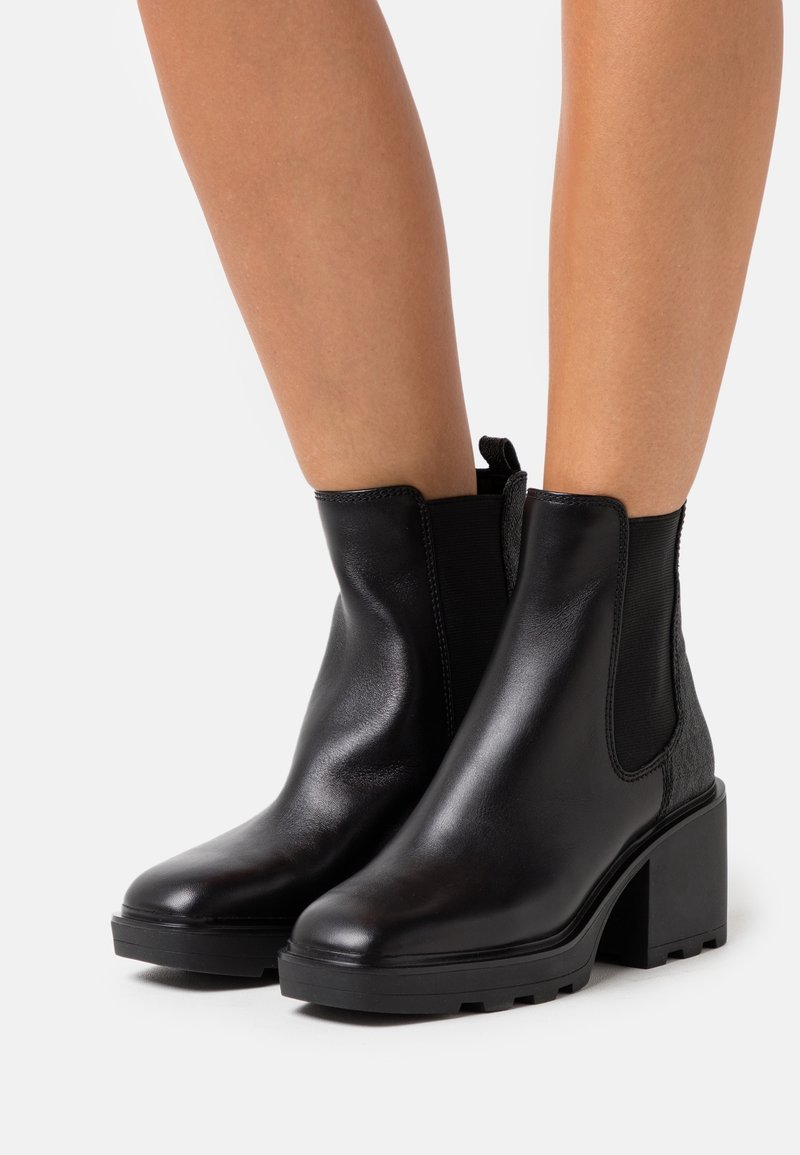 Bottes basses en cuir noir avec talons carrés, panneaux latéraux élastiques et accents texturés à l'arrière. Finition lisse, bout rond, et semelle robuste.