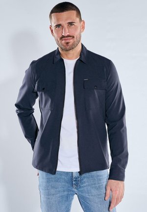 Mann mit kurzen Haaren und Bart, der eine marineblaue Jacke mit Reißverschluss über einem weißen Hemd und hellblauen Jeans trägt, vor einem einfarbigen Hintergrund steht.