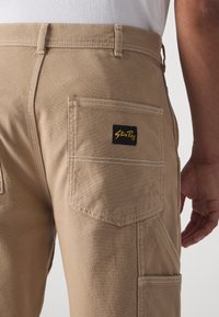 Tan trousers made from durable fabric. Features a patch pocket with a black label and white stitching accents. Classic workwear design.