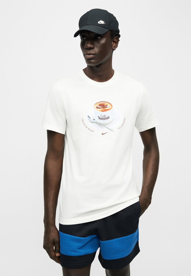 Giovane uomo con indosso una maglietta bianca con una tazza di caffè e la scritta "Café Swoosh", pantaloncini neri e blu, e un cappellino Nike nero, in piedi con una mano in tasca.