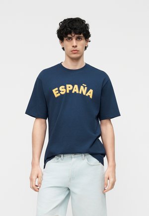 Młody mężczyzna o kręconych ciemnych włosach, ubrany w granatowy t-shirt z napisem "ESPAÑA" żółtymi literami i jasnoniebieskie szorty jeansowe.
