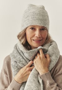 Grijze gebreide muts en sjaal gemaakt van zacht, gestructureerd materiaal. De sjaal hangt losjes om de nek en toont een gemengd grijs patroon.
