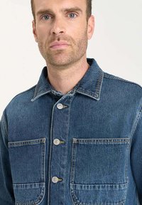 Donkerblauwe denimjas met een knoopsluiting aan de voorkant, puntige kraag, twee borstzakken en contrasterende stiksels.