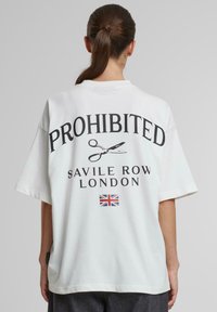 Valge üle mõõtude T-särk, millel on kiri "KEELATUD" ja käärid graafika, all on "SAVILE ROW LONDON" ning UK lipu aktsent.