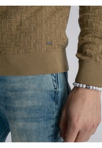 Khaki gebreide trui met een textuurpatroon, geribbelde manchetten en een klein logo accent, gecombineerd met lichtblauwe denim jeans en een kralenarmband.