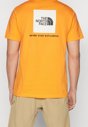Personne portant un t-shirt orange avec le logo "THE NORTH FACE" et le texte "NEVER STOP EXPLORING" dans le dos, assorti à un pantalon beige.