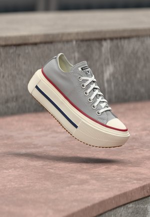 Grauer Sneaker mit niedrigem Schaft, weißen Schnürsenkeln, dicker cremefarbener Sohle, roten und blauen Streifen, schwebt über einer Steinformation im Freien.