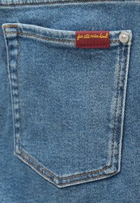 Calças de ganga azul claro com um bolso traseiro retangular. Apresenta uma etiqueta bordô com "for all mankind" em costura amarela.