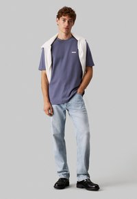 T-shirt grigio a maniche corte, collo rotondo, con un piccolo logo; maglione bianco appoggiato sulle spalle; jeans blu sbiaditi; scarpe nere.