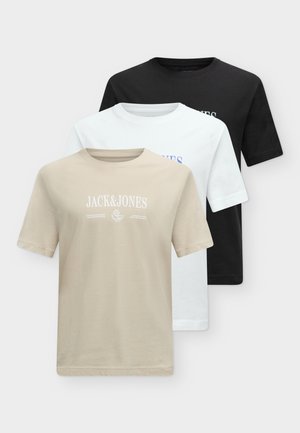 Mladý muž v čiernom tričku Jack & Jones, voľných modrých džínsoch a čiernych topánkach stojaci s jednou rukou v vrecku na bielom pozadí.