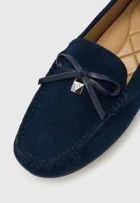 Mocassini in camoscio blu navy con punta arrotondata, caratterizzati da un fiocco in pelle e un piccolo ciondolo metallico. Interno strutturato con cuciture a motivo diamantato.