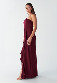 Bordna enodelna obleka z nagubanim volanom in prilagojenim zgornjim delom, opremljena s stranskim razporkom za lažje gibanje.