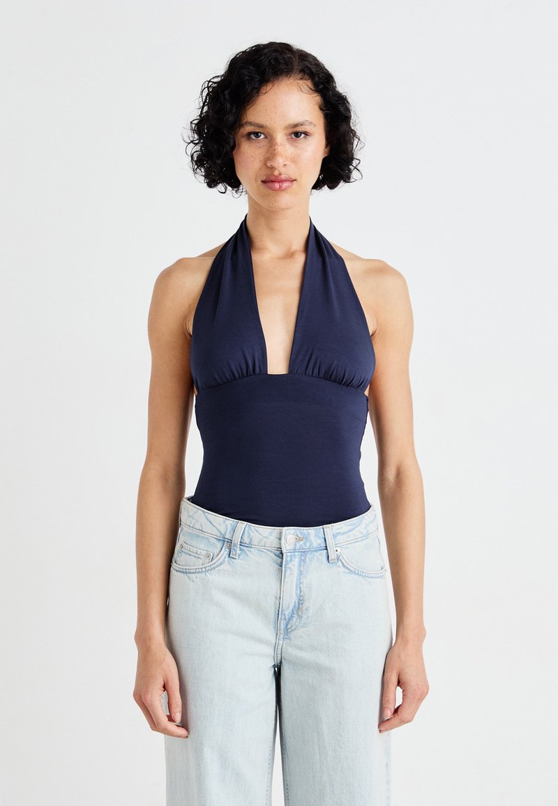 Navy halter neck bodysuit with deep V front, smooth fabric, and fitted design, paired with light blue high-waisted jeans.