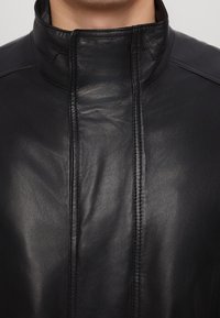 Sort læderjakke med høj krave, glat tekstur og synlige syninger langs forstykkets sømme. Ingen knapper eller metaldele synlige.