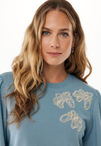 Světle modrá mikina s třemi texturovanými, vyšitými květinovými motivy zdobenými korálky na přední straně. Hladká tkanina s žebrovaným výstřihem.