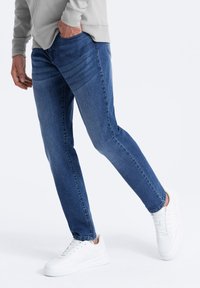 Siniset farkut, joissa on kapea leikkaus ja hienovaraiset kulutuskohdat edessä, yhdistettynä valkoisiin tennareihin. SuUrisen kankaan pinta.