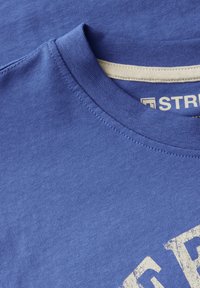 Niebieska bawełniana koszulka z okrągłym dekoltem, krótkimi rękawami i jasnobeżowymi szwami. Na przodzie znajduje się blady biały napis w wyraźnej czcionce.