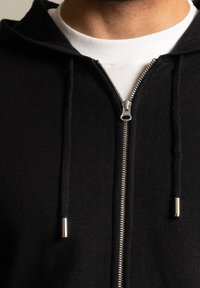 Sudadera con cremallera negra de acabado texturizado, con una cremallera plateada, cordones y una camiseta de cuello redondo blanca debajo.
