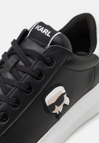 Zwarte leren sneaker met ronde neus, platte veters en rubberen zool. Heeft een cartoonembleem en "KARL" merktekens op de tong.
