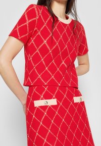La mujer lleva un top de punto rojo y una falda con un patrón de diamantes beige y pequeñas perlas incrustadas, un cuello beige y acentos de bolsillos abotonados.