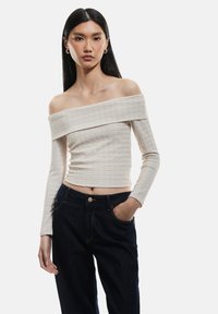 Paita slim-fit ilman olkapäitä, valmistettu tekstuurista beigeistä kankaasta, yhdistettynä korkeavyötäröisiin tummansinisiin farkkuihin. Pitkät hihat, lyhyt malli.