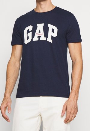 Muž nosí tmavě modré tričko s krátkým rukávem s velkými bílými písmeny "GAP" a malým červeným nápisem "Original", doplněné o bílé kalhoty.