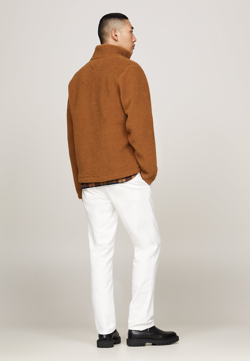 Giacca in pile marrone con colletto alto, tessuto texturizzato e vestibilità rilassata. Abbinata a pantaloni bianchi e stivali neri con tacco.