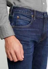 Granatowe dżinsy mają slim fit z delikatnym połyskiem, pięć kieszeni, złoteHardware i kontrastowe pomarańczowe przeszycia.