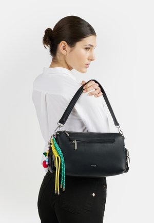 Donna con capelli scuri raccolti in uno chignon indossa una camicia bianca e pantaloni neri, tenendo una borsa nera decorata con nappine colorate sulla spalla.