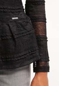Haut noir texturé en dentelle à manches longues avec volants superposés et petit logo Guess en métal, porté avec un pantalon gris clair.