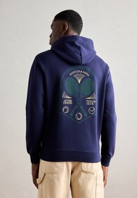 Haut à capuche bleu marine avec un motif de raquettes de tennis et de balles au dos, avec des accents verts et jaunes. Matière en mélange de coton, coupe décontractée.
