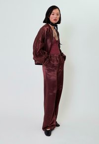 Burgundi aksomo bomber klasikinė striukė su elastingomis rankovėmis, derinama su atitinkama aksomo palaidine ir plačiomis kelnėmis; stiliuota su juodu chokeriu.