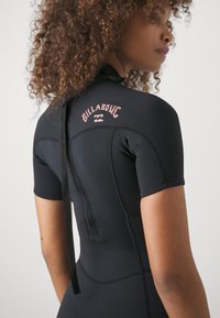 Une combinaison de plongée en néoprène noir à manches courtes, avec le logo rose "Billabong" au dos et une attache dans le dos. Texture lisse et design ajusté.