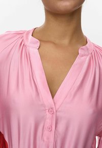Pink button-up blouse with a V-neckline, featuring soft fabric, pleated detailing, and a contrasting accent on the left side.