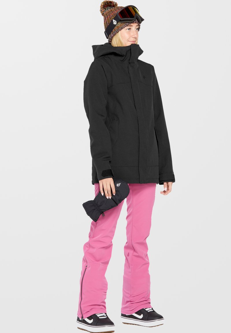 Zwarte skijas met een hoge kraag, roze sneeuwbroeken en zwarte wanten. Het model draagt een meerkleurige gebreide muts en een skibril, op zwarte sneeuwlaarzen.