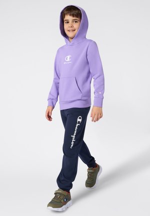 Sweat-shirt à capuche violet avec poche avant et logo blanc. Pantalons de jogging bleu marine avec le texte blanc "Champion" sur le côté. Baskets grises et vertes.