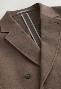 Gros plan d’un blazer marron texturé avec des revers crantés, un seul bouton visible et une bordure grise à l’intérieur du col.
