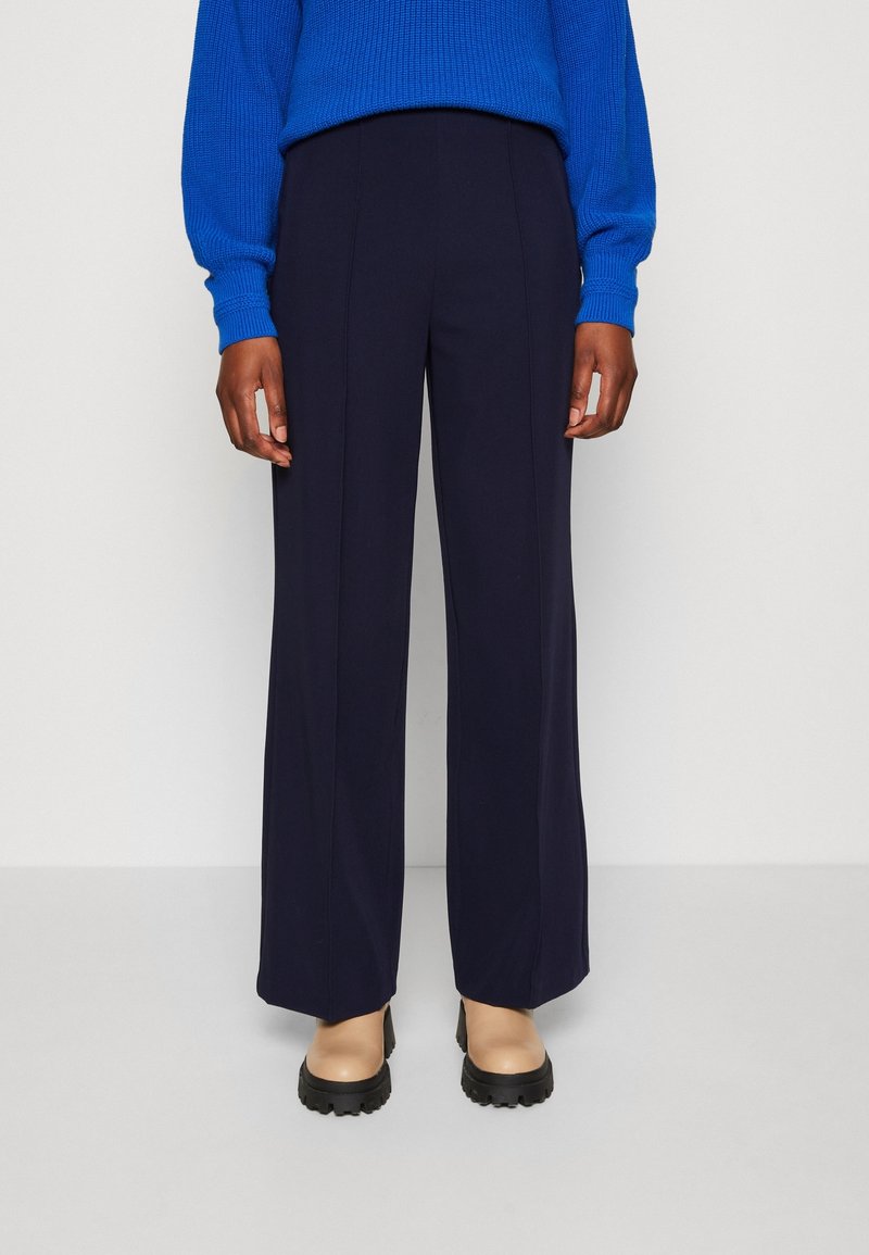Pantalon large bleu marine avec une texture lisse et une coupe ajustée, associé à des chaussures beiges à plateforme et un pull texturé bleu vif.