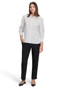 Witte blouse met lange mouwen en knoopsluiting, versierd met zilveren studs op de borst; gecombineerd met zwarte broek en zwarte blokhakken.