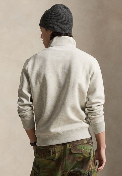Man met een lichtgrijze trui, camouflagebroek en een donkergrijze gebreide muts, met de rug naar de camera tegen een effen beige achtergrond.