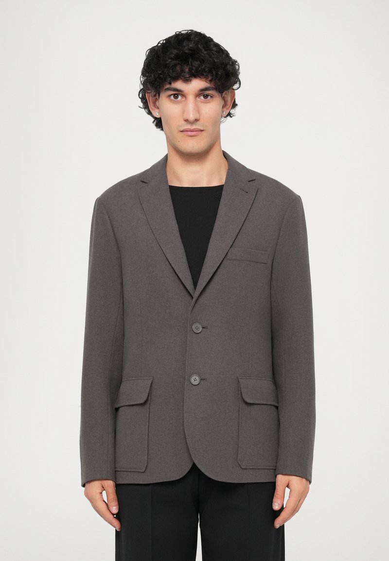 Blazer cinza com dois botões, lapelas notched e um único bolso no peito. Apresenta dois bolsos frontais de remendo e uma textura suave.
